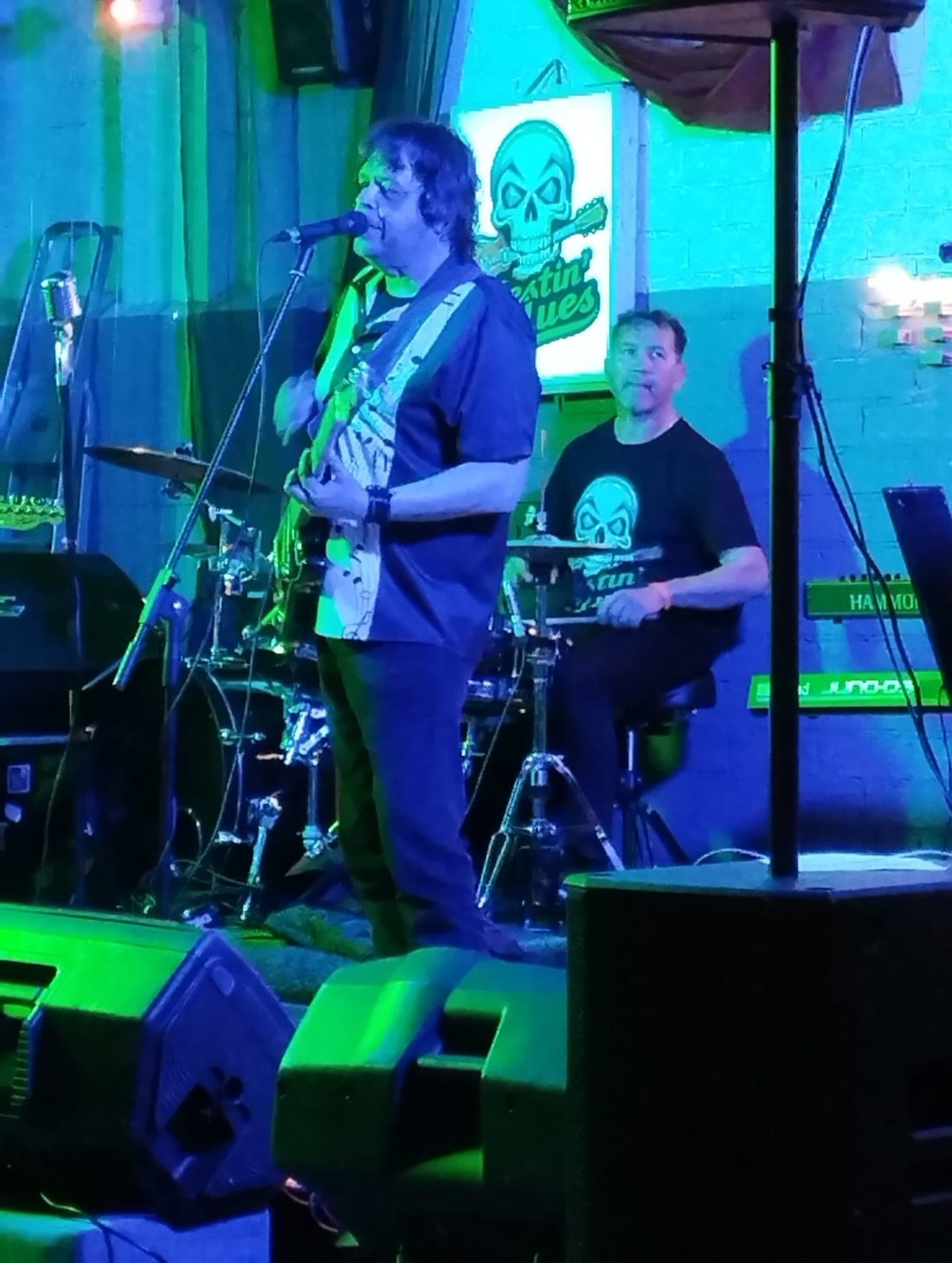 Isi cantando y Alberto a la batería, bajo luz azul y verde, con el cartel de la banda detrás